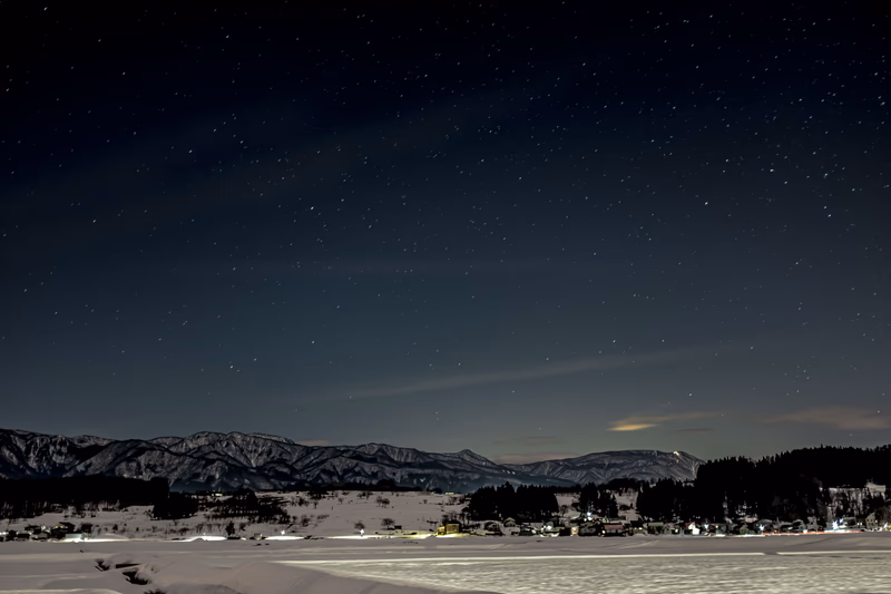 A serene winter night with snow-covered mountains and a lake reflecting the stars above. The scene is illuminated by the soft glow of distant lights.