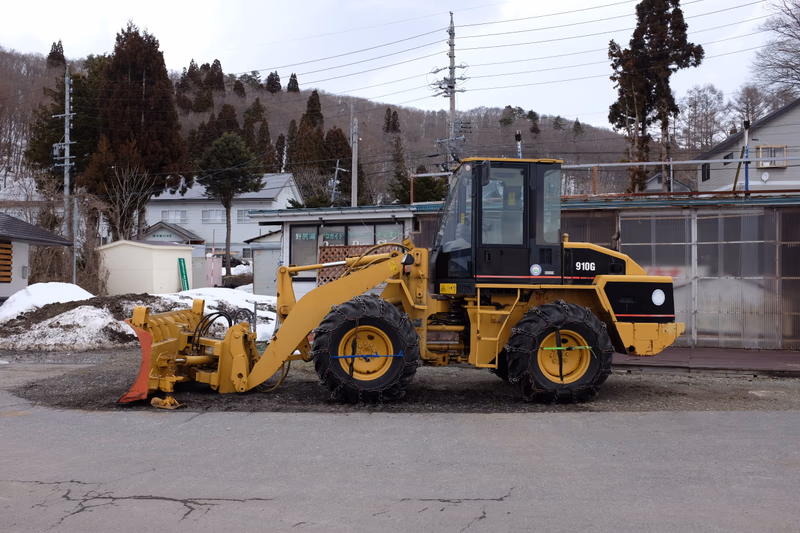 A yellow front loader with a snowplow attachment is parked in a residential area with houses and trees in the background.