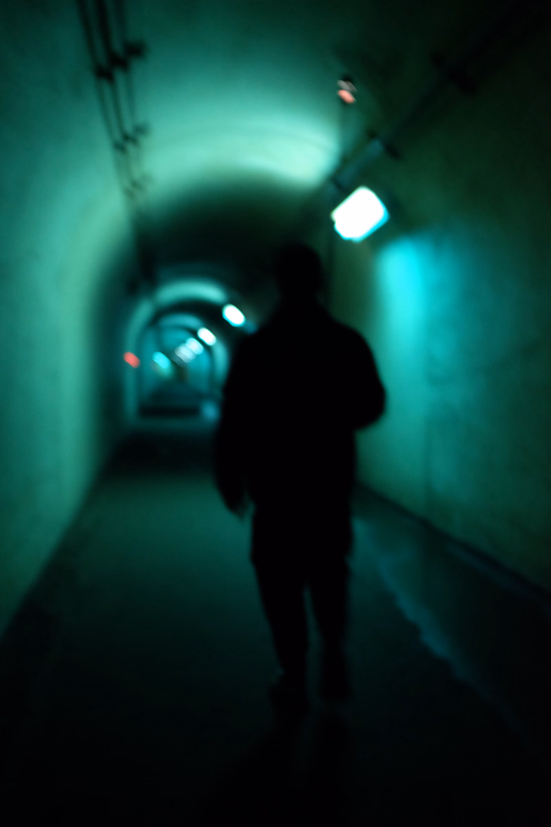 A silhouette of a person walks down a brightly lit, curved tunnel or hallway with overhead lights.