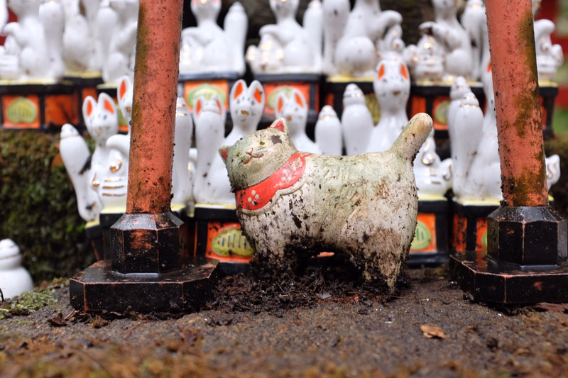 A small, weathered ceramic figurine sits on the dirt ground next to the base of a wooden beach hut post, with many white statues in the background.