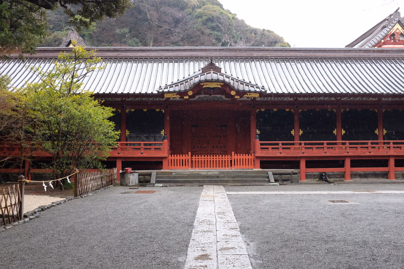 A traditional Japanese temple building with a dark tiled roof stands in front of a backdrop of green mountains.