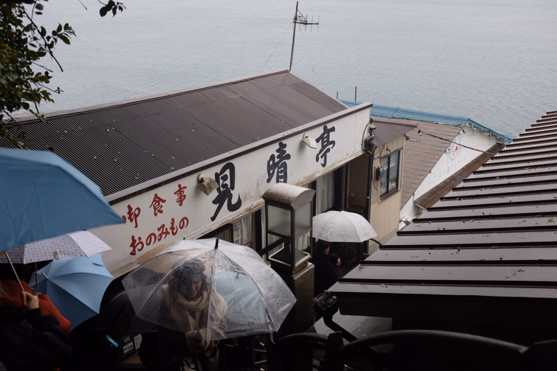 A restaurant with a sign reading "御食事 見晴亭 おのみもの" overlooks the sea, with several umbrellas in the foreground.