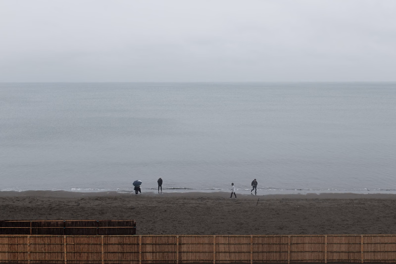 Several people are walking along the sandy beach near the ocean under an overcast sky, with a wooden fence in the foreground.