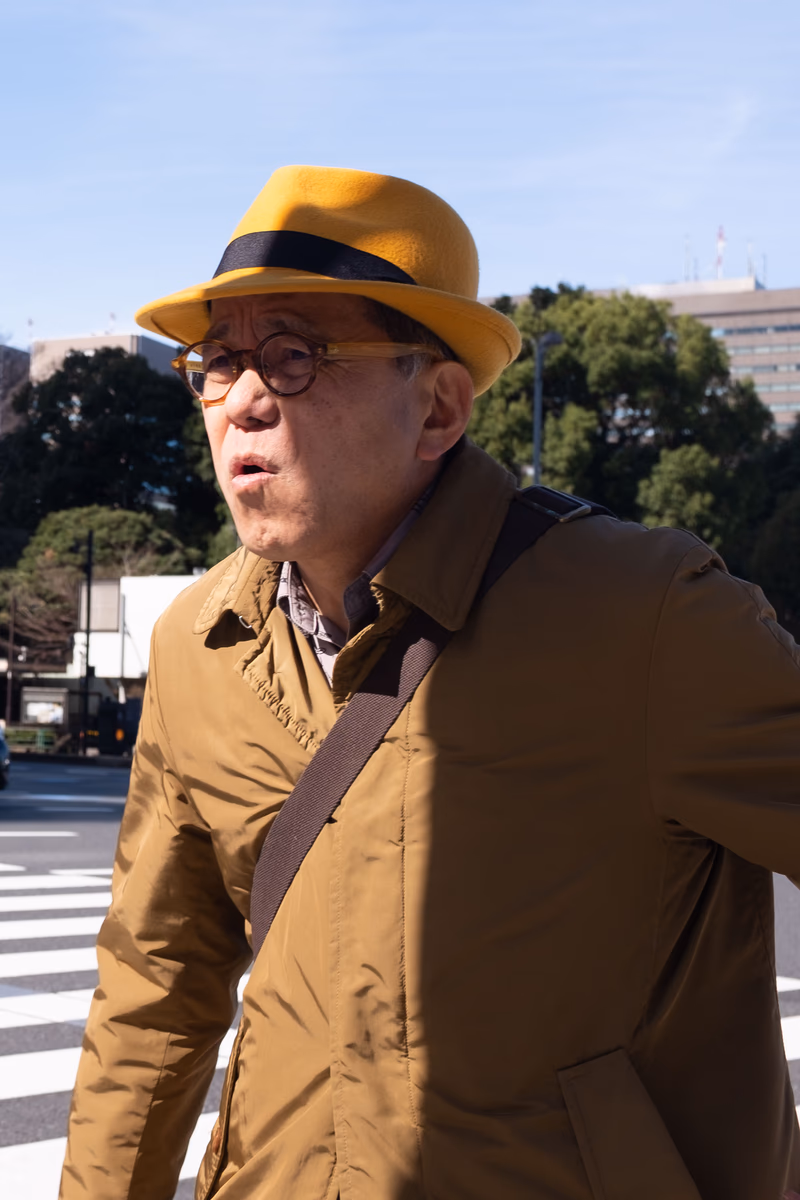 A man wearing a yellow hat and a brown jacket, standing in front of a crosswalk.
