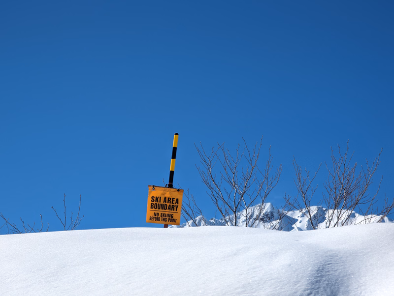 A yellow and black sign reading "SKI AREA BOUNDARY NO SKIING BEYOND THIS POINT" stands in the snow under a clear blue sky.