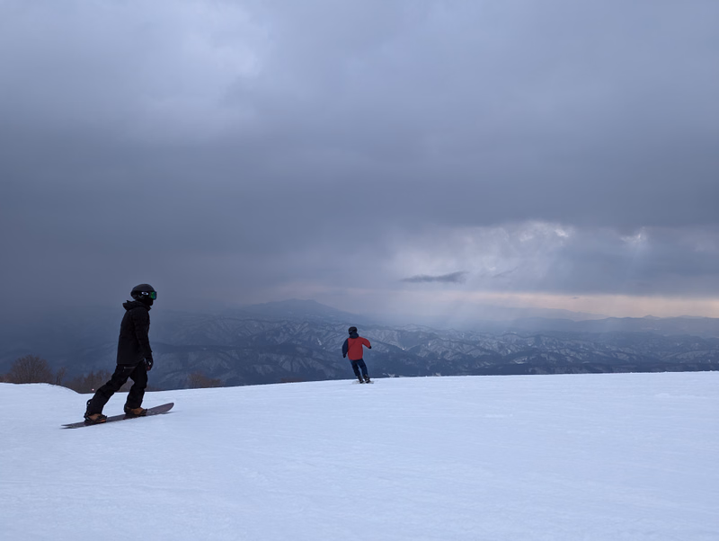 Two snowboarders are visible on a snowy slope under a heavily overcast, gray sky overlooking distant mountains.