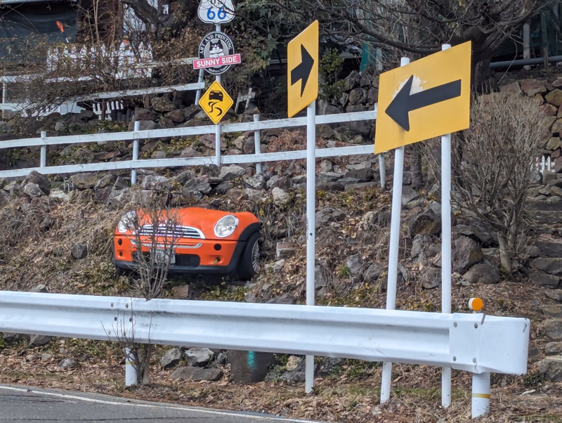 An orange classic car is parked near a white fence with directional signs, one reading "SUNNY SIDE" and another showing a black arrow.