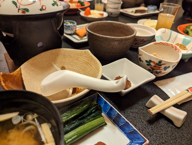 A collection of small bowls, a black teapot, and chopsticks are arranged on a dark textured table, suggesting a traditional Japanese meal.