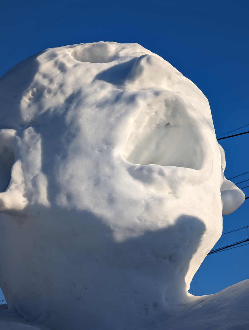 A large, white snowman sculpture with carved-out facial features is photographed against a clear blue sky.
