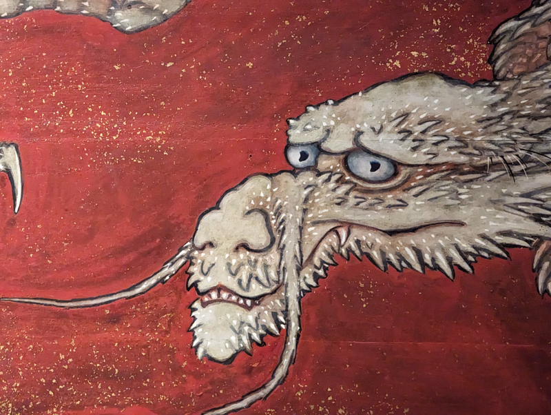 A detailed painting of a grotesque, hairy monster with sharp teeth and wide, fearful eyes against a red background.