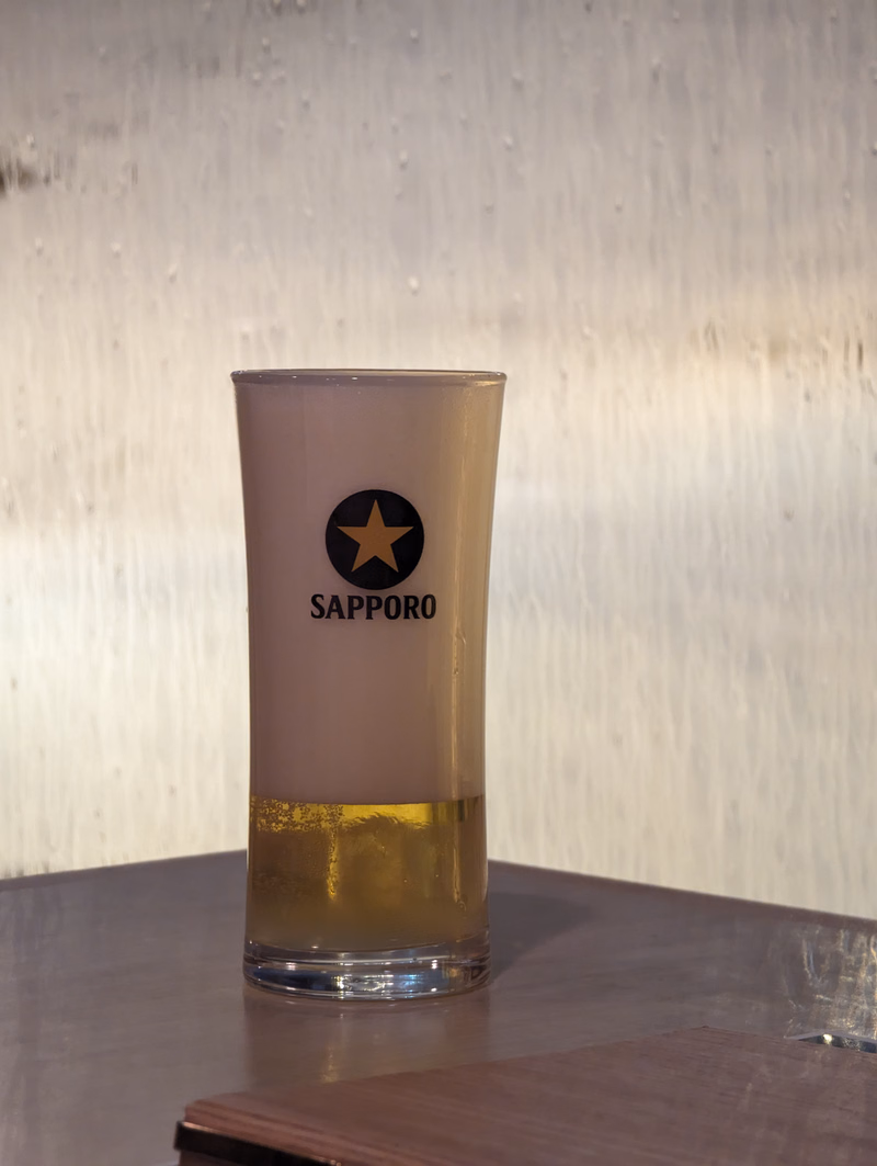 A tall glass of Sapporo beer sits on a dark surface, featuring the Sapporo logo.