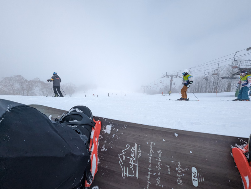 A snowboarder is visible on a snowy slope under a cloudy sky, with ski lifts in the background.