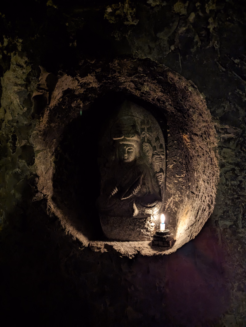 A dark, mossy cave niche houses a stone statue illuminated by a small candle.