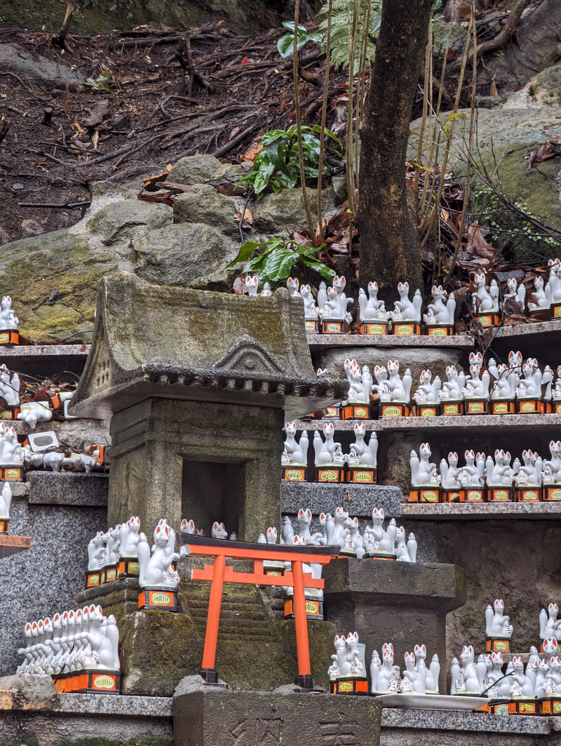 A tiered stone structure adorned with numerous white statues and a small red torii gate, set against a backdrop of mossy rocks and dense foliage.