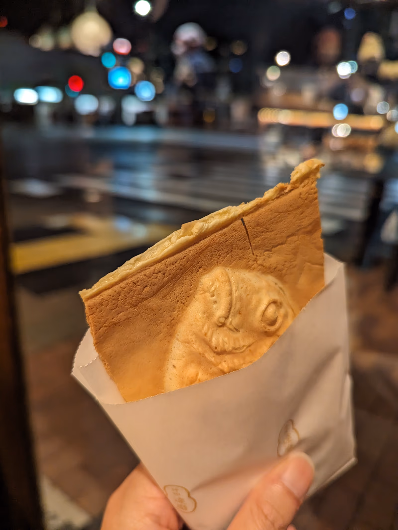 A close-up of a fish-shaped waffle in a paper bag, held by a hand in front of a blurred city street at night.