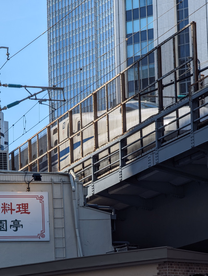 An elevated train structure passes in front of modern glass skyscrapers, with a sign reading "米料理 岡亭" visible below.
