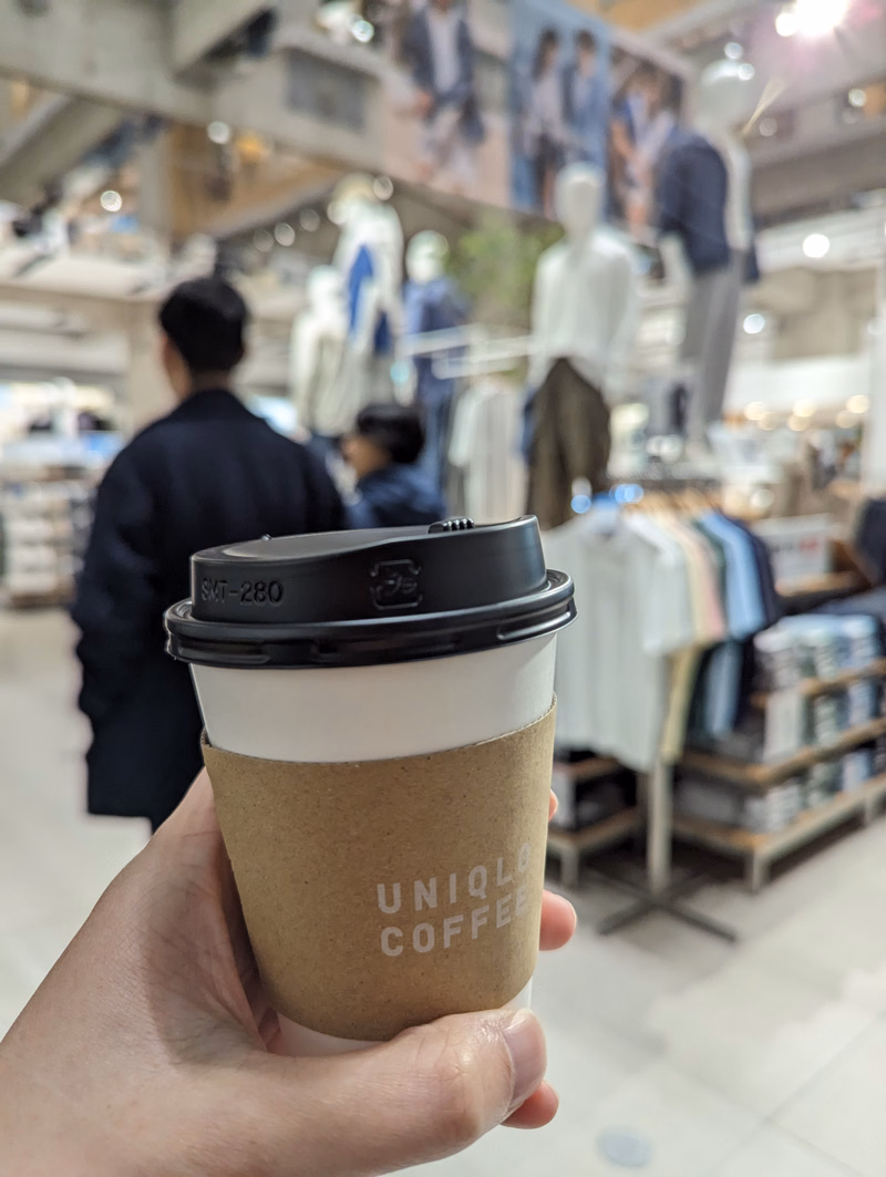 A hand holds a brown paper coffee cup labeled "UNIQLO COFFEE" inside a brightly lit retail store.