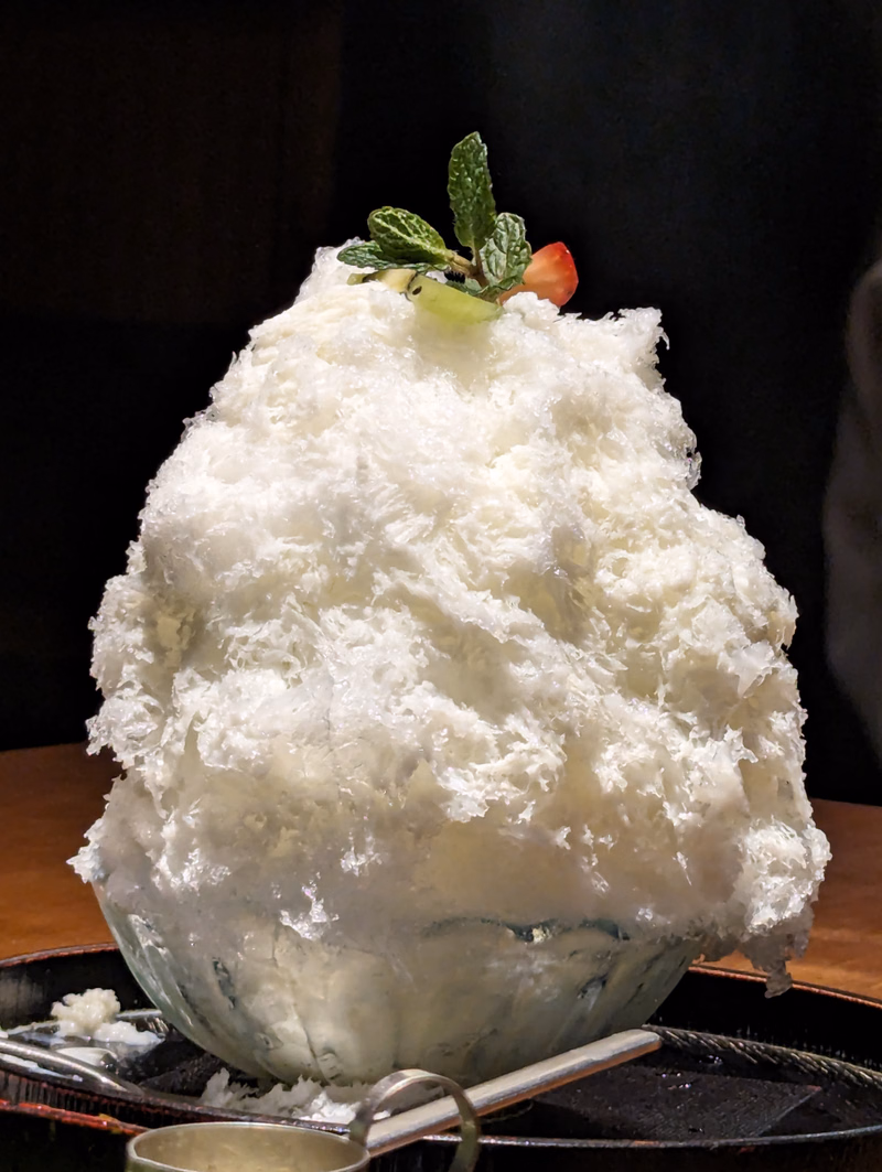 A large mound of white coconut dessert is topped with fresh mint leaves and a strawberry, served on a dark platter.