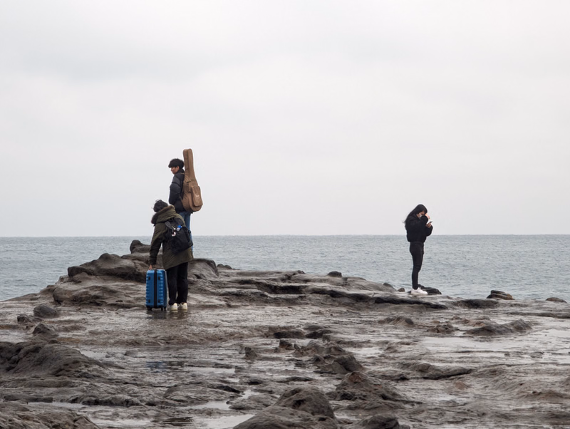 Several people are standing on dark, wet rocks along the coast overlooking the ocean under an overcast sky.