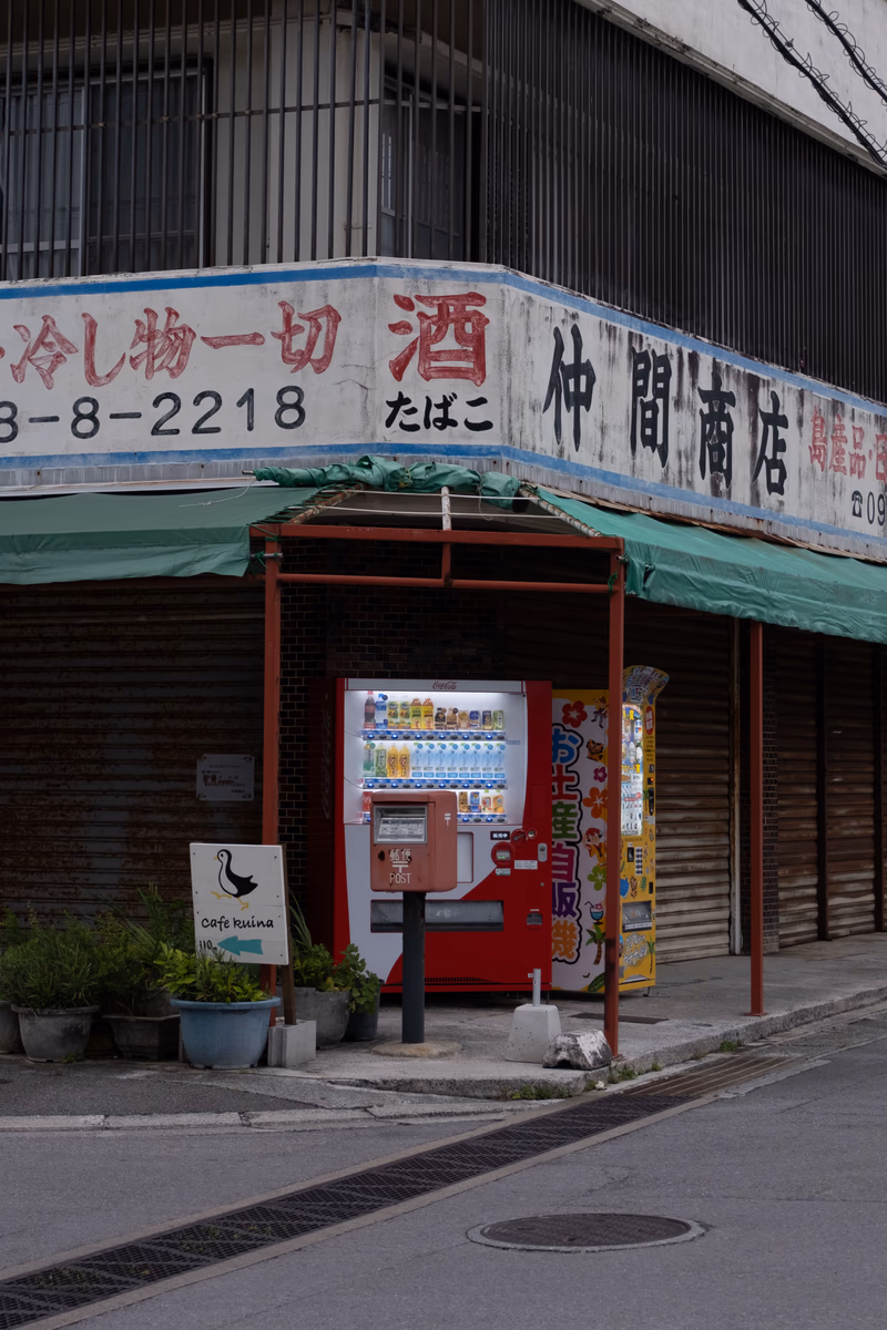 A street scene in Ishigaki, Okinawa, Japan, featuring a vending machine, a small shop, and a few potted plants.