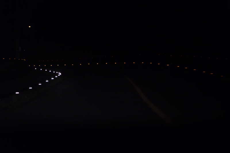 A dark night scene showing a road illuminated by numerous small lights stretching into the distance.