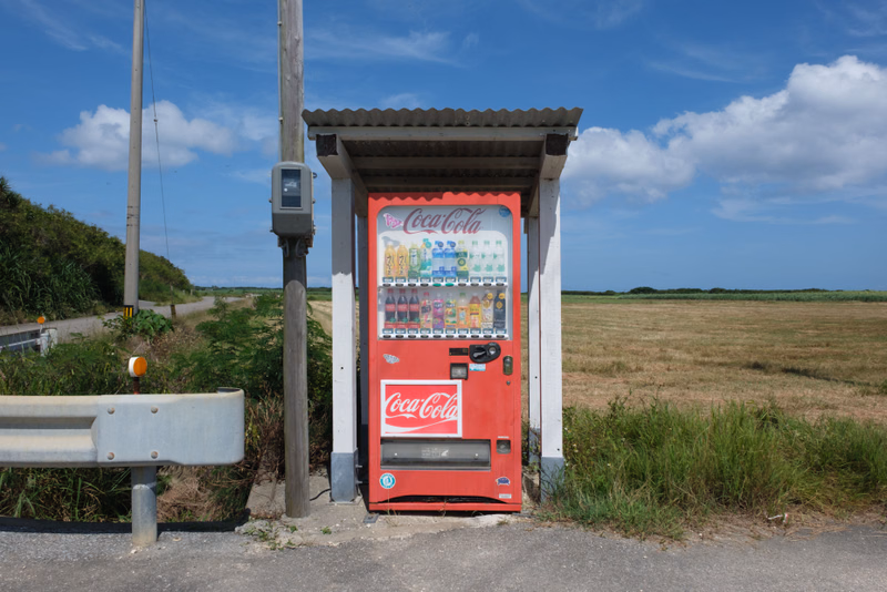 A red Coca-Cola vending machine stands outdoors near a grassy field under a blue sky with white clouds.