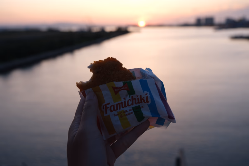 A person holding a piece of food with a wrapper that says 'Famichioki' near a sunset and a body of water.