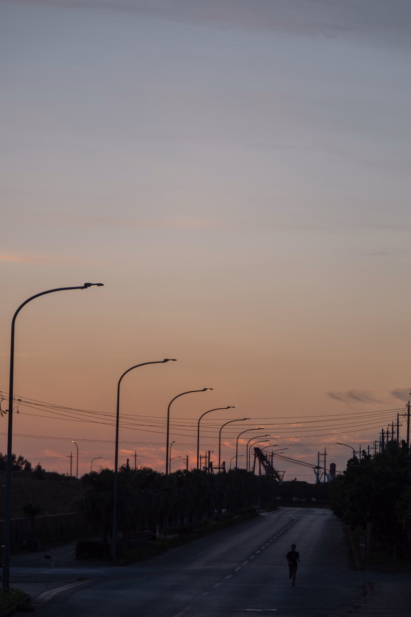A long, straight road stretches into the distance under a vast, pale orange and pink sunset sky, lined with utility poles.
