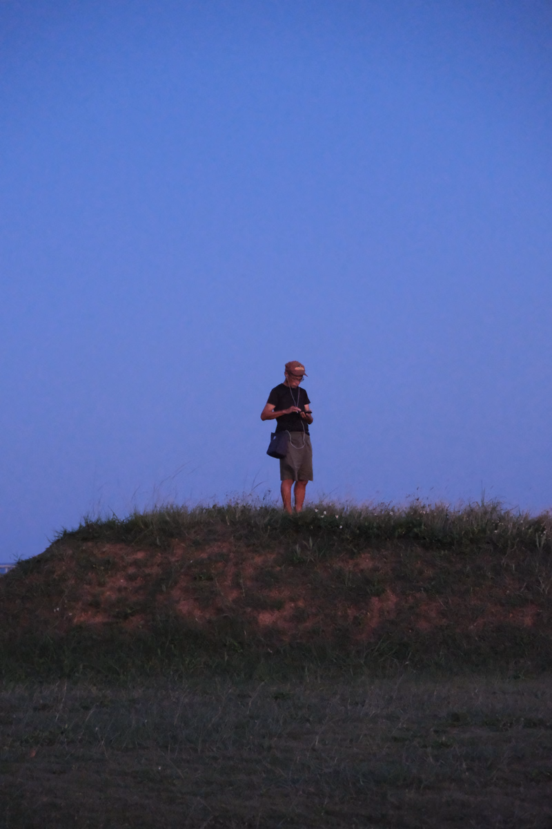 A person stands on a grassy hill against a clear blue sky, holding a small object.