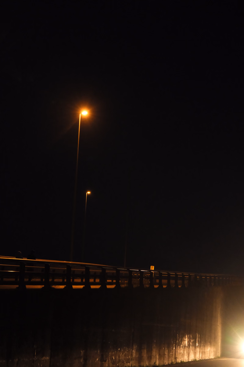 A street light illuminates a bridge at night, with another distant light visible in the dark sky.
