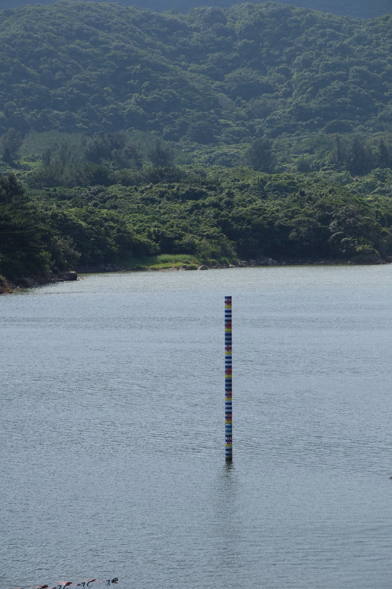 A colorful vertical marker stands in the calm water of a lake, backed by lush green, forested mountains.