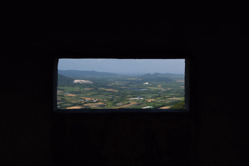A panoramic view of a green countryside valley with scattered fields and distant hills under a bright sky.