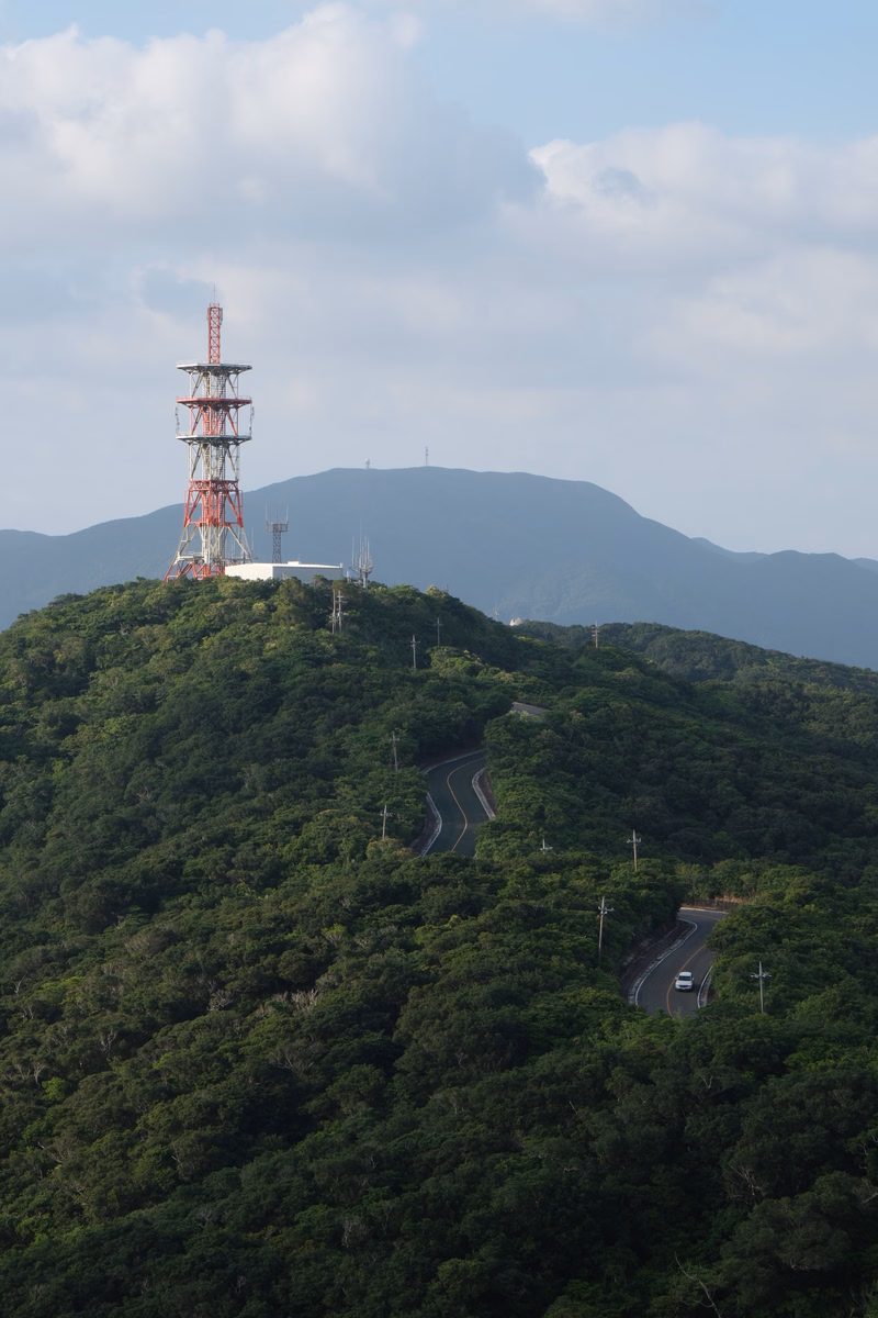 A tall cell tower stands on a forested hill overlooking winding roads in a mountainous landscape under a cloudy sky.