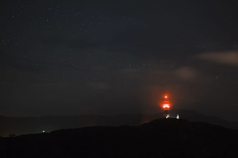 A red lighthouse shines brightly on a dark hill against a starry night sky over the sea.