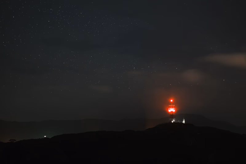 A nighttime photograph of a mountain range with a light tower in the distance.