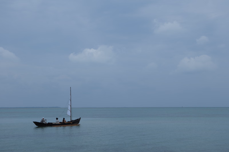 A small traditional boat with a mast is visible on the calm ocean under a cloudy sky.