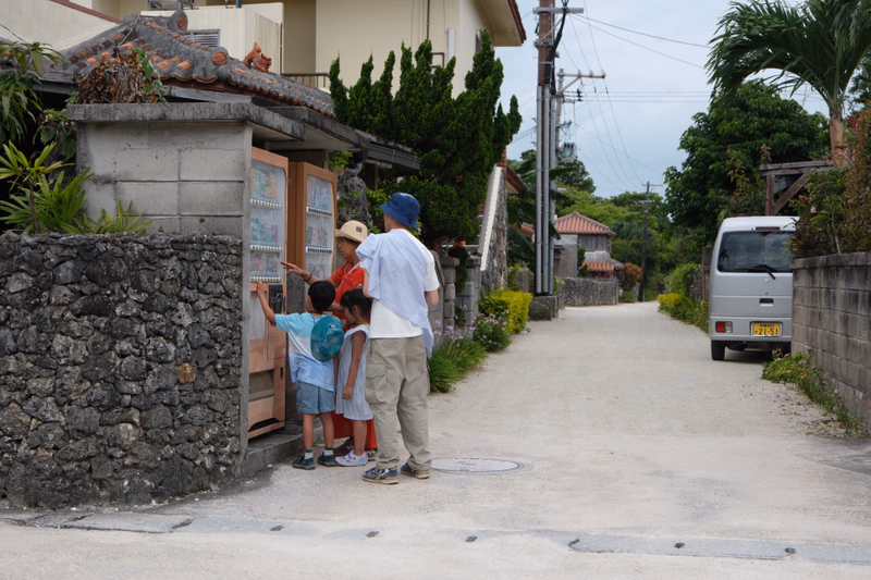 Several children stand near a small building on a paved street with a white van parked further down the road.