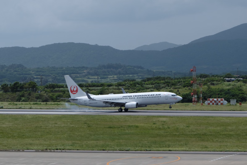 A Japan Transocean Air airplane is visible on the runway under an overcast sky.