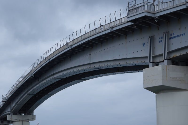 A large concrete viaduct with guardrails spans across the frame under a cloudy sky, featuring Japanese text that reads "沿岸部事務所施設".
