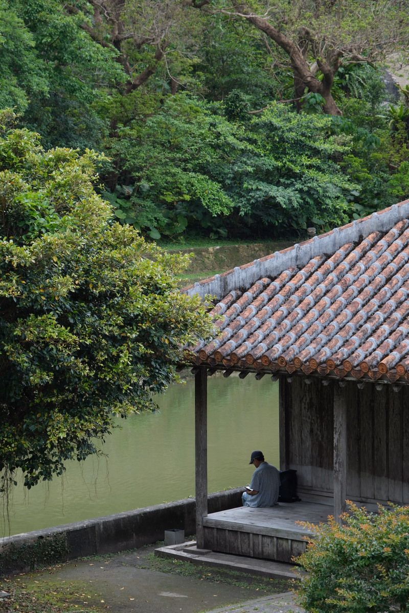 A person sits under a tiled roof hut overlooking a calm pond surrounded by lush green trees.