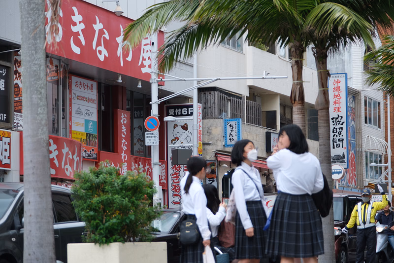 Several people walk on a street lined with storefronts, including one with red signage that reads "きたな屋" and another with "おさな屋".