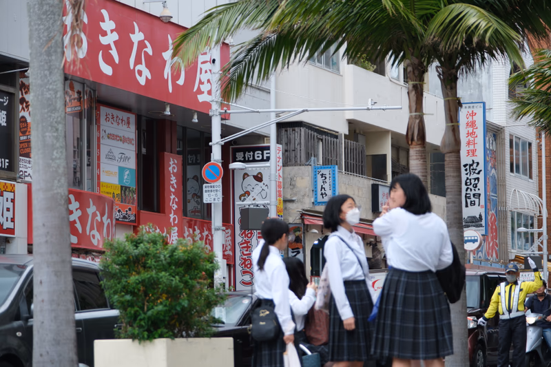 A street scene in Naha, Okinawa, Japan, featuring a group of people in school uniforms and a variety of colorful signs.