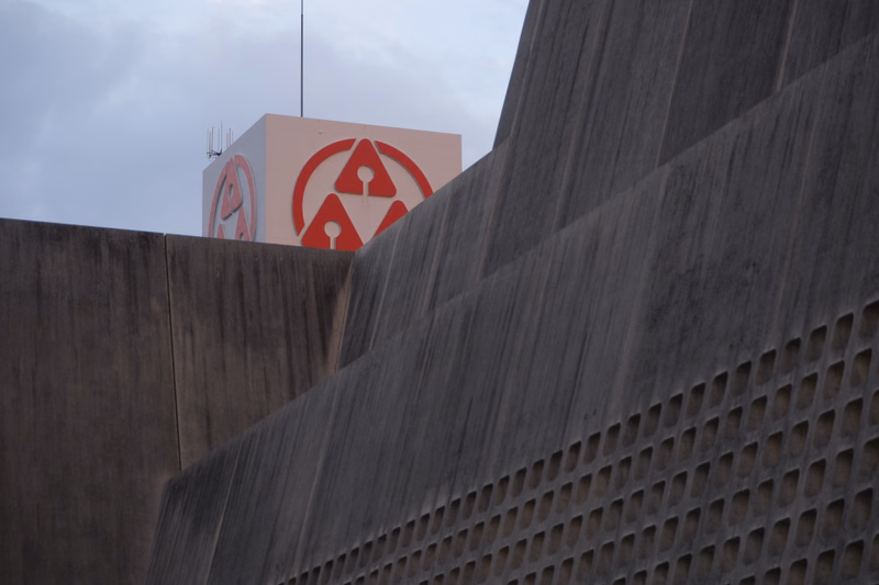 A large, textured concrete wall dominates the foreground, with a white structure featuring a red circular logo visible behind it under a cloudy sky.