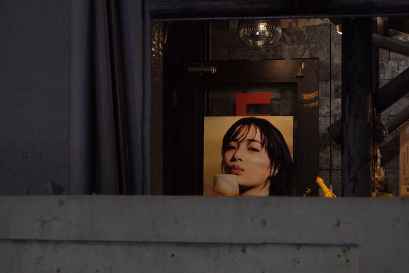 A poster of a woman is visible through a dark storefront window, with a red 'E' sign above it.