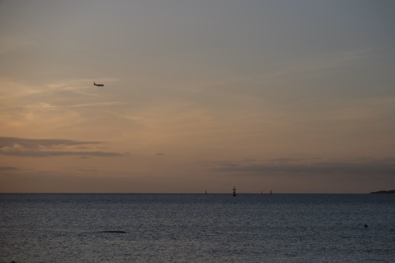A small airplane flies over a calm sea under a pale, hazy sky near sunset.