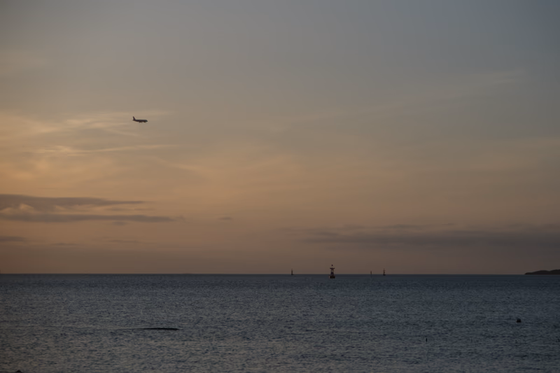 A serene seascape at sunset with a plane flying low over the ocean and a lighthouse in the distance.