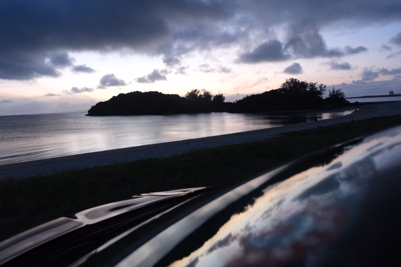 A car parked near a small island with a calm sea and a cloudy sky