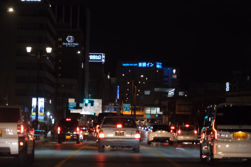 A nighttime view of a busy city street with traffic, featuring a DoubleTree hotel building and signs like "toyoko-inn.com".