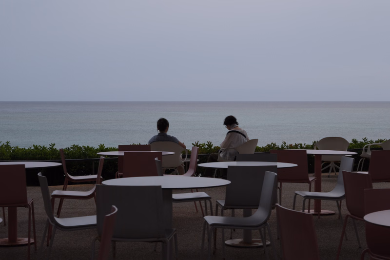 Two people sit at a table overlooking the calm sea on a cloudy day, surrounded by outdoor seating.