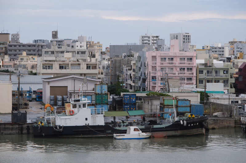 A fishing boat is docked in a harbor area surrounded by dense urban buildings under a cloudy sky.
